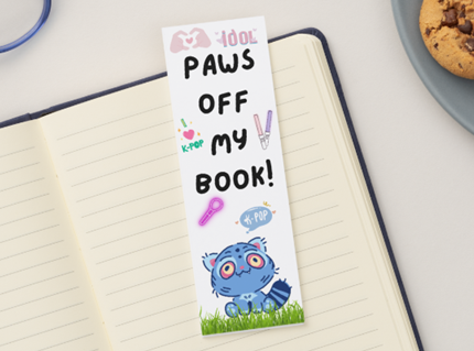 Bookmark with cartoon cat design and text 'Paws Off My Book!' on a notebook with cookies and glasses in the background.